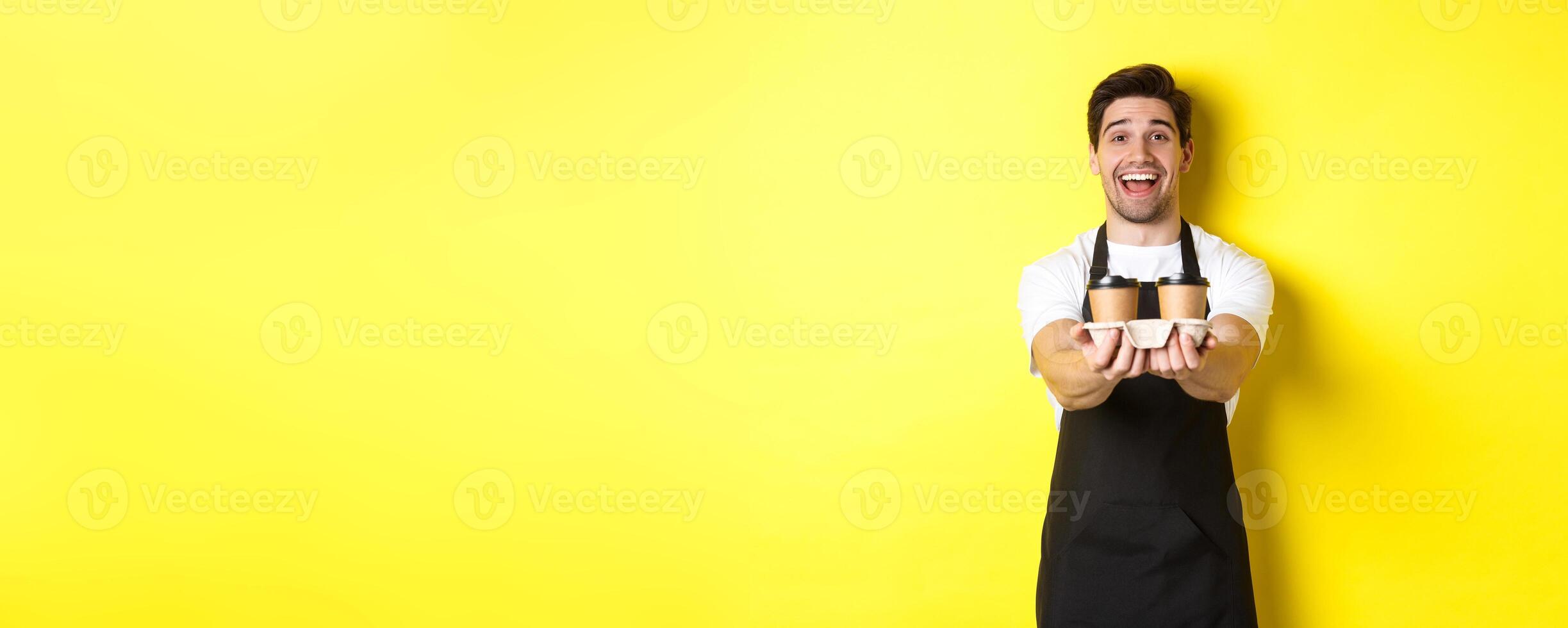 Friendly barista in black apron giving takeaway order, holding two cups of coffee and smiling, standing over yellow background photo