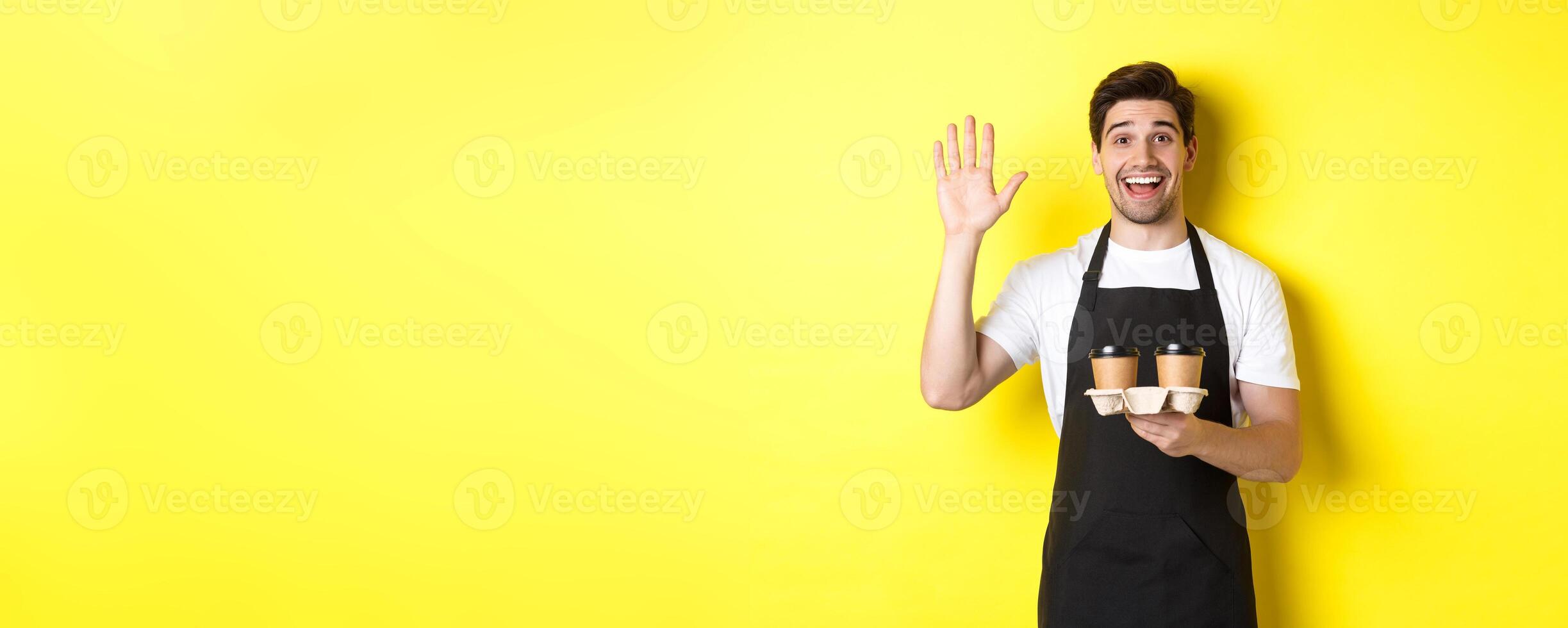Friendly waiter in cafe waving hand at customer, holding takeaway coffee oder, standing against yellow background in black apron photo