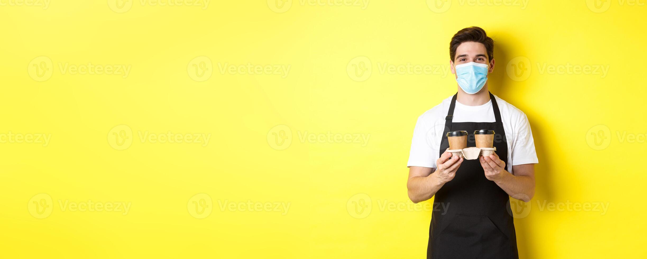 Concept of covid-19, cafe and social distancing. Barista in medical mask serving coffee in takeaway cups, standing in black apron against yellow background photo