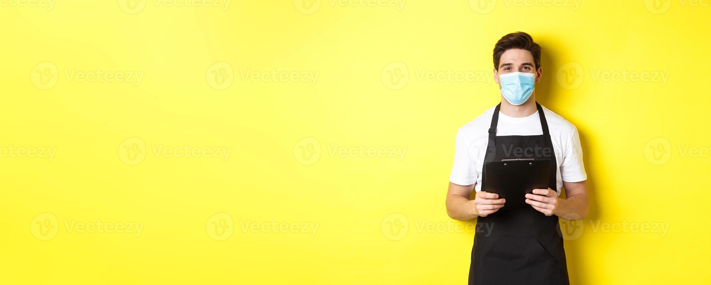 Concept of covid-19, small business and quarantine. Salesman in black apron and medical mask holding clipboard, working in shop, standing over yellow background photo