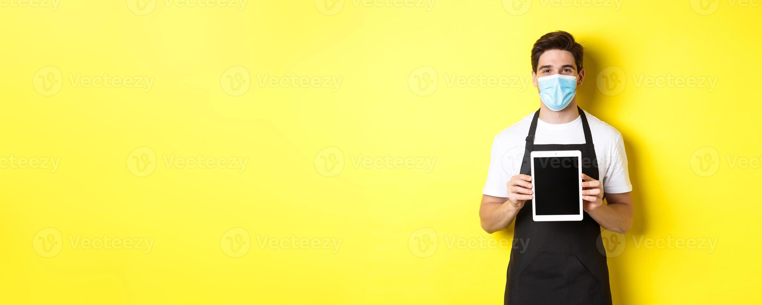 Concept of covid-19, small business and pandemic. Smiling barista in medical mask showing tablet screen, standing over yellow background photo
