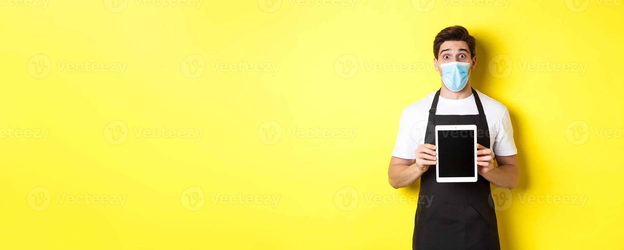 Concept of covid-19, small business and pandemic. Surprised waiter in black apron and medical mask showing tablet screen, standing over yellow background photo