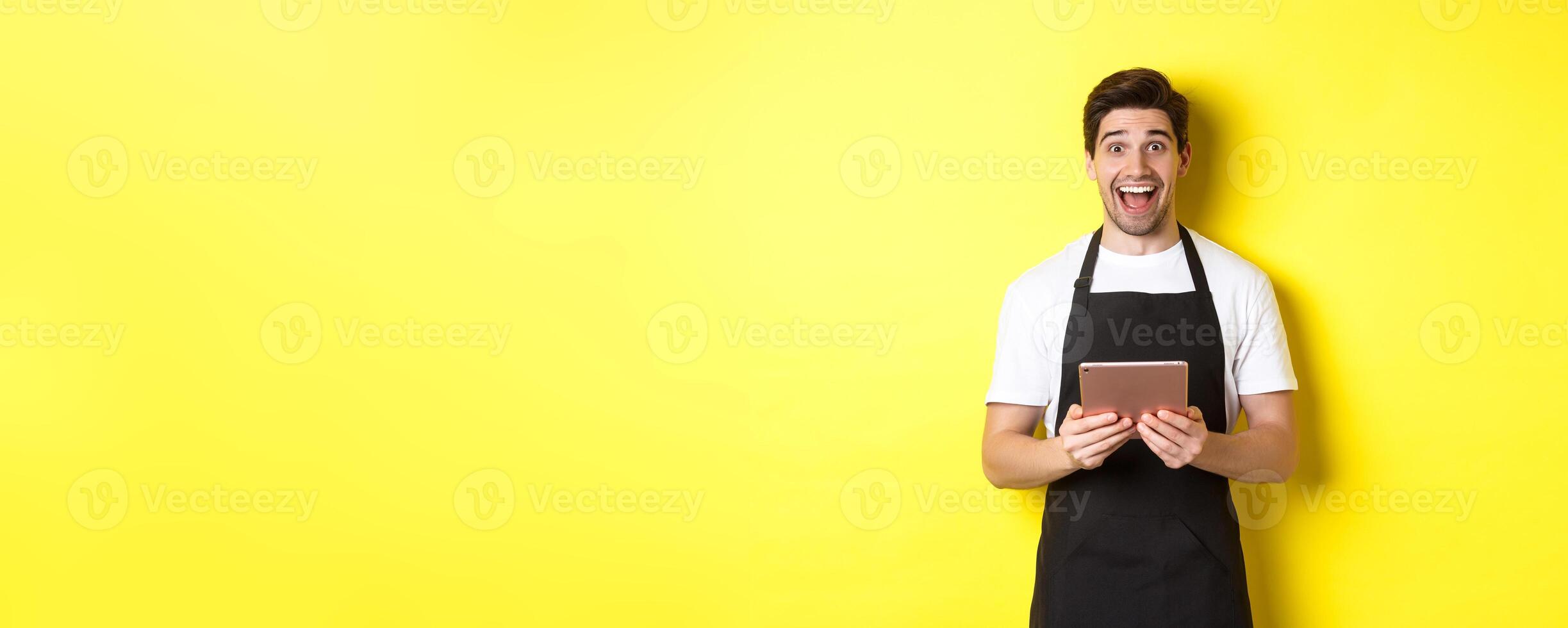 Happy salesman in black apron, holding digital tablet and looking surprised, standing against yellow background photo