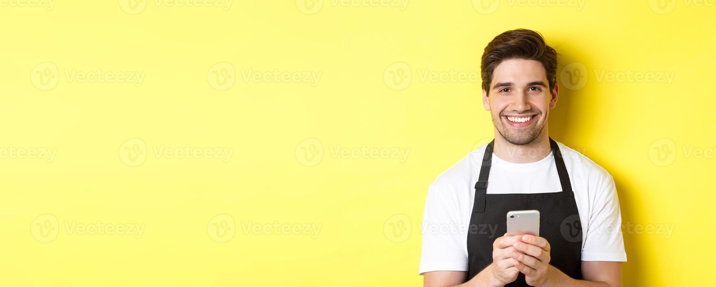 Close-up of handsome barista sending message on mobile phone, smiling happy, standing over yellow background photo