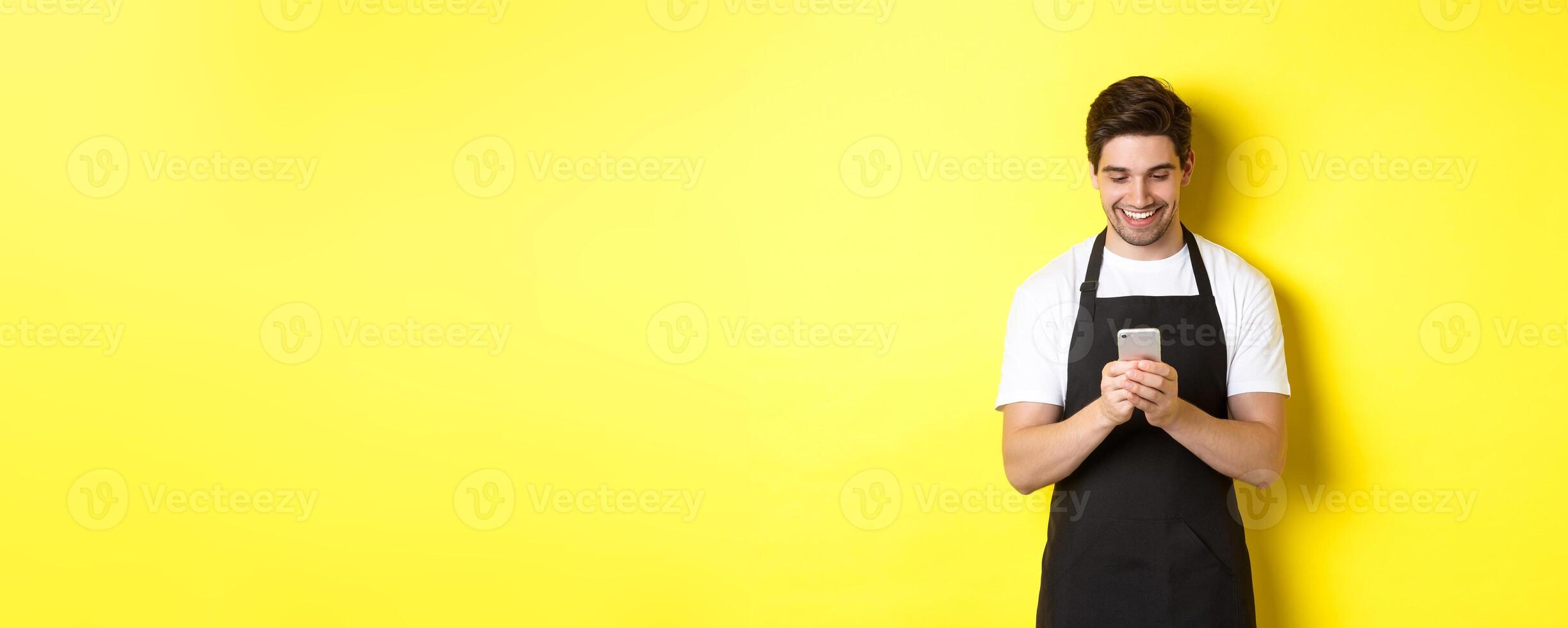 Waiter in black apron reading message on mobile phone, smiling happy, standing over yellow background photo