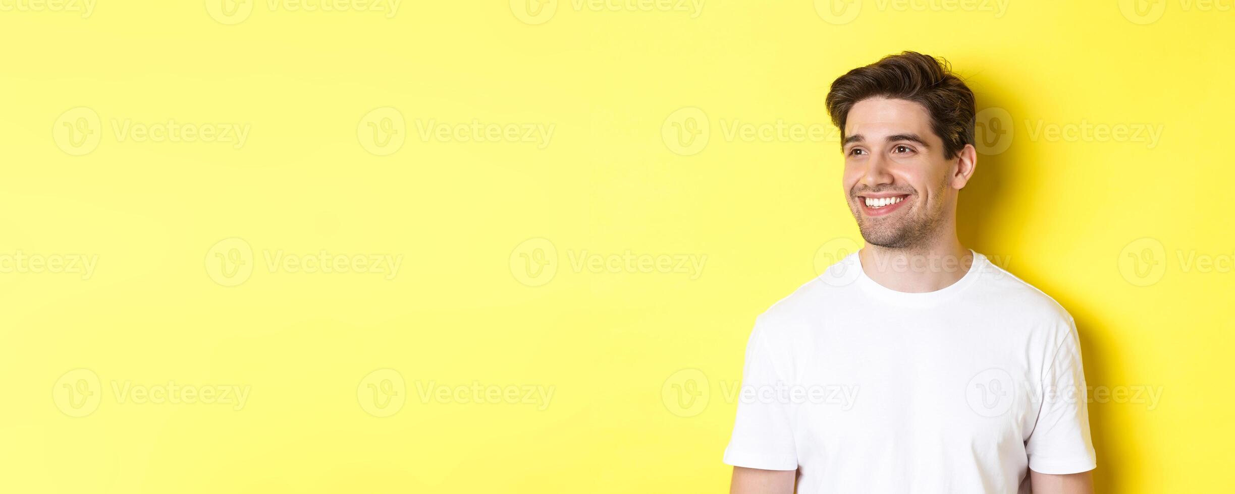 Close-up of attractive bearded man in white t-shirt smiling, looking left at copy space, standing against yellow background photo