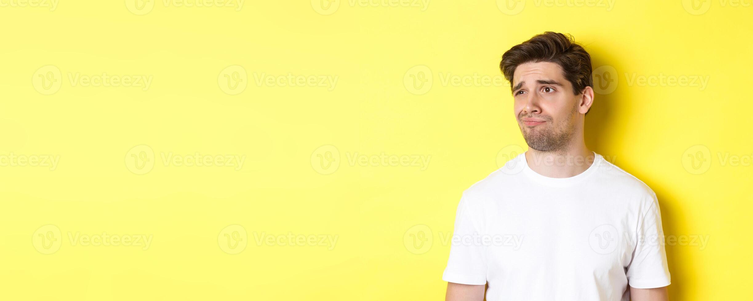 Reluctant guy in white t-shirt looking left, grimacing skeptical and displeased, standing over yellow background photo