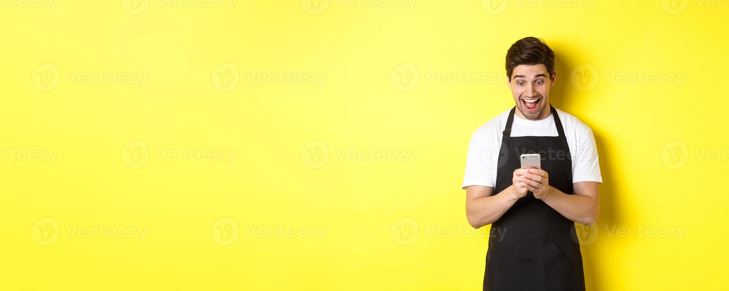 Barista looking surprised as reading message on mobile phone, standing in black apron against yellow background photo