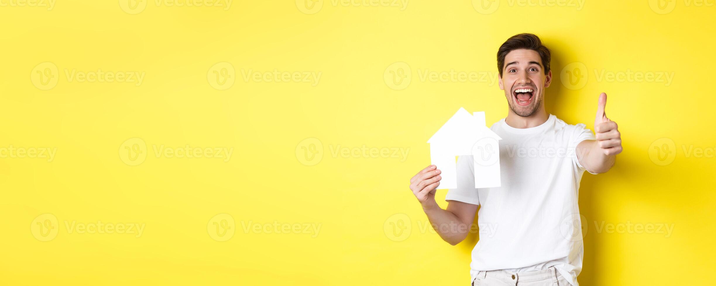Real estate concept. Happy young male buyer showing thumb up and paper house model, smiling satisfied, standing over yellow background photo