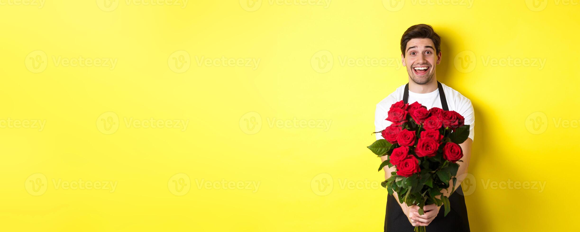 Seller in flower shop wearing black apron, giving bouquet of roses and smiling, standing over yellow background photo
