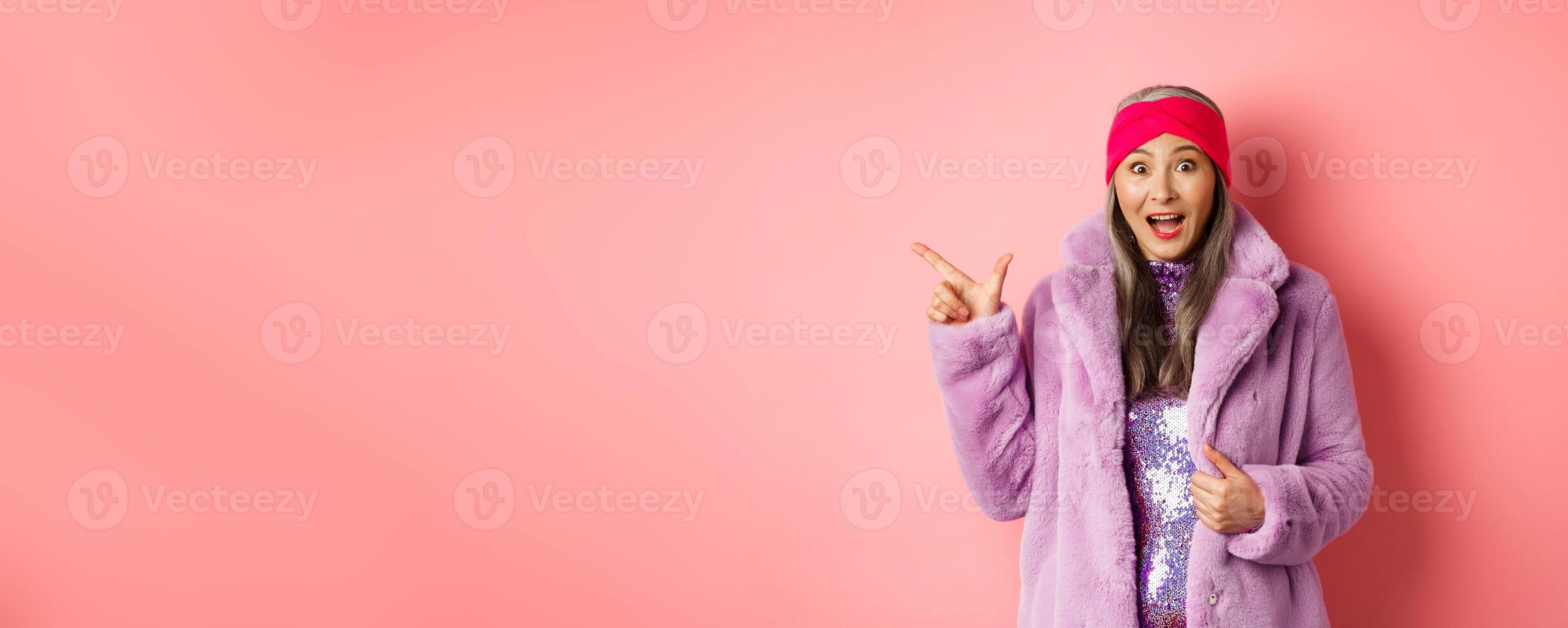 Fashion and shopping concept. Fashionable senior asian woman in winter coat and headaband pointing finger left at copy space, staring amazed at camera, checking out banner photo