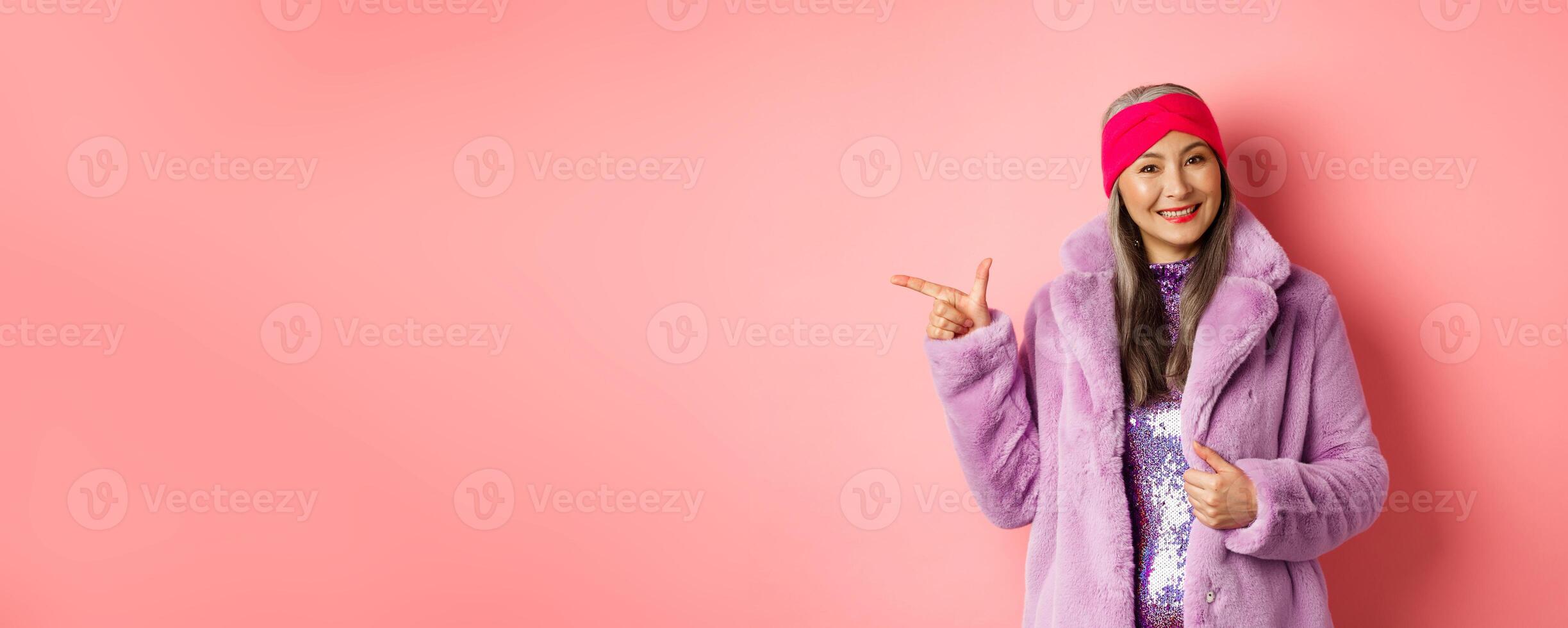 Fashion and shopping concept. Stylish senior asian woman pointing finger left at promotion deal, smiling at camera, suggesting special offer, standing against pink background photo