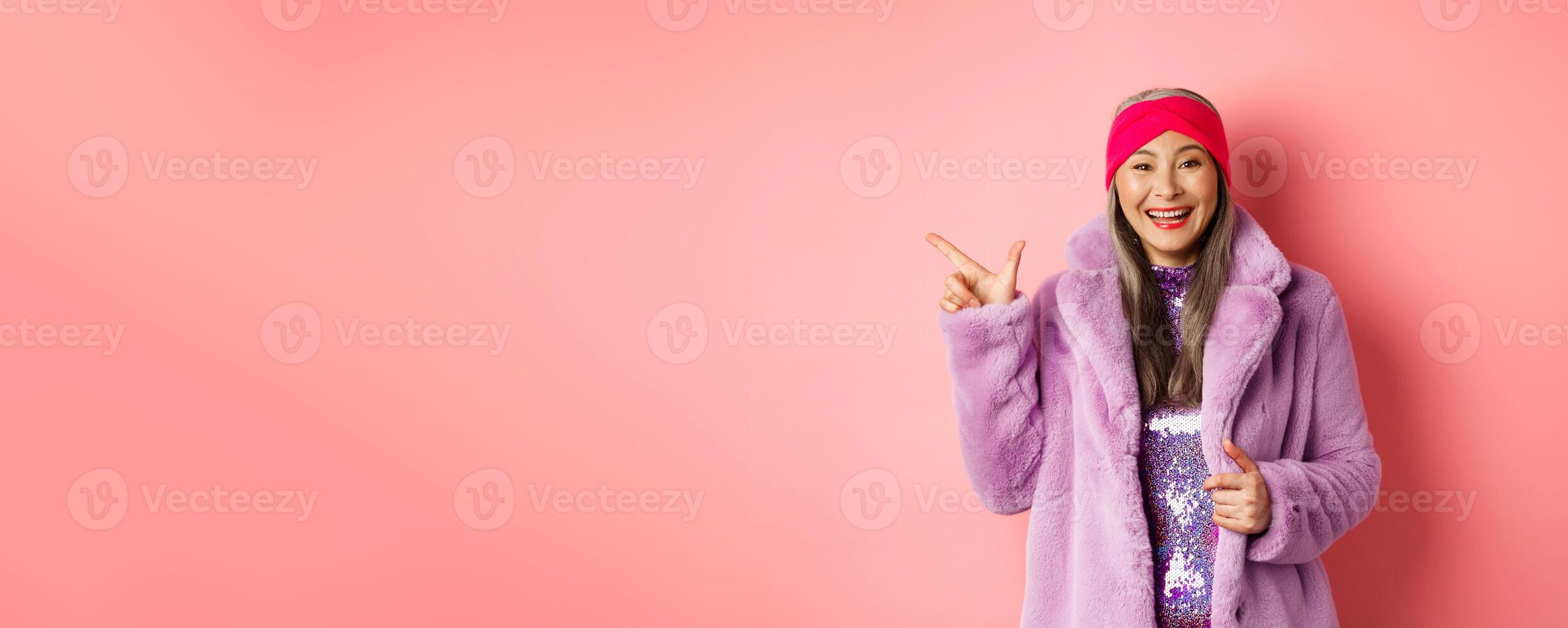 Fashion and shopping concept. Happy asian senior woman checking out trendy promotion, pointing finger left at logo and smiling, wearing winter fake fur coat and headband photo