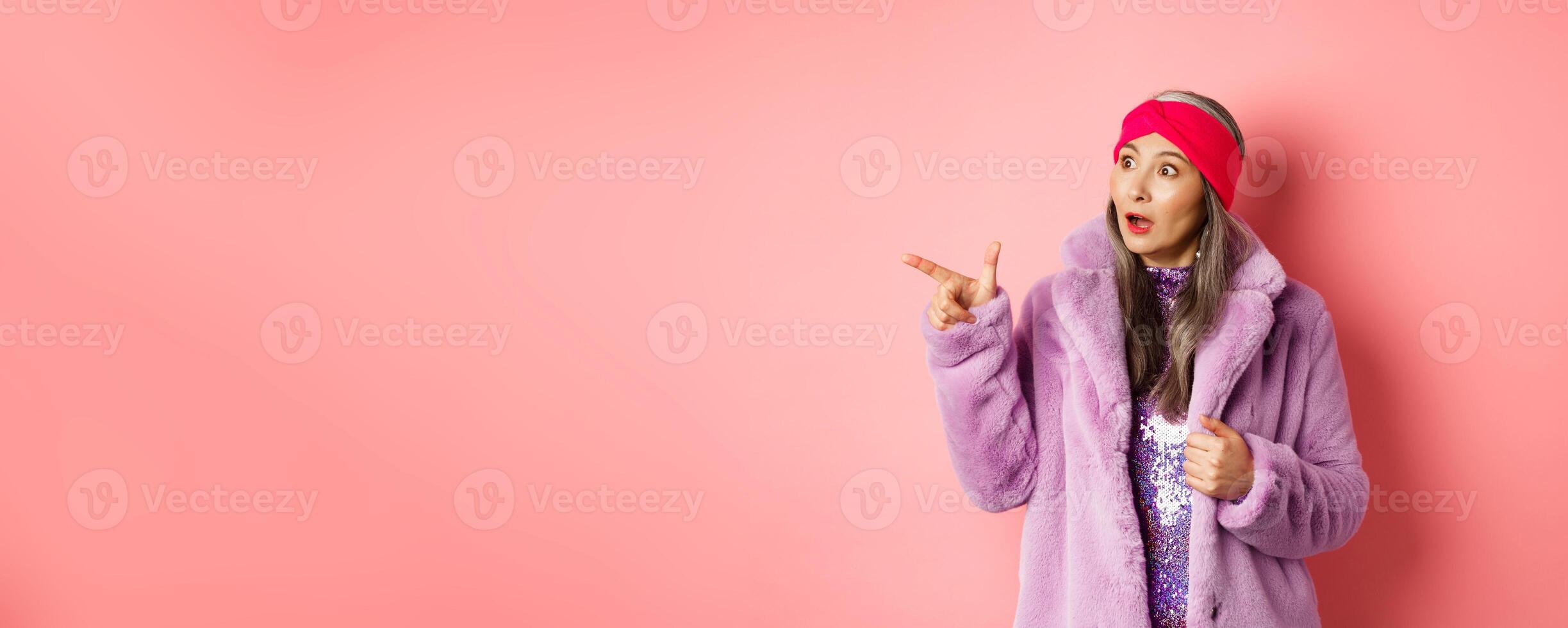 Fashion and shopping concept. Trendy elderly asian female in fake fur and glittering dress pointing, looking left with amazement, checking out offer, pink background photo