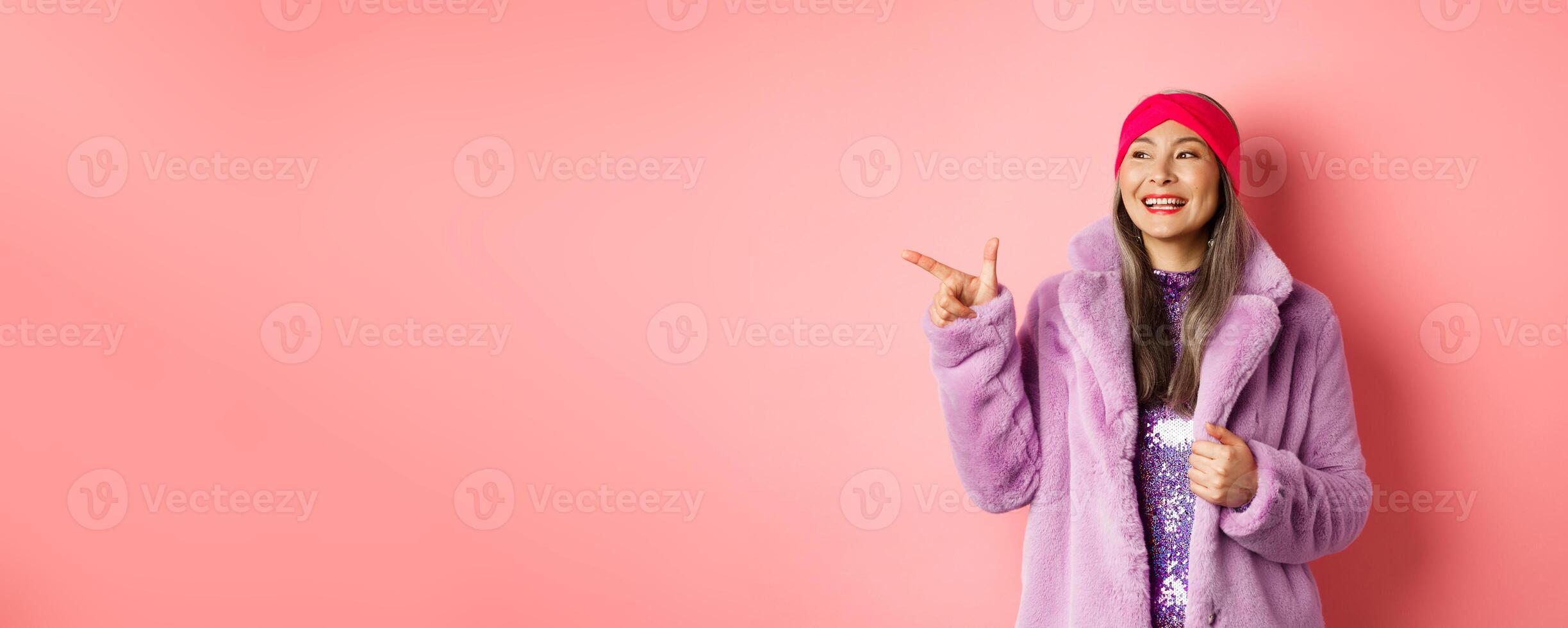Happy senior asian woman smiling, pointing finger left and looking satisfied, checking out special promotion, pink background photo