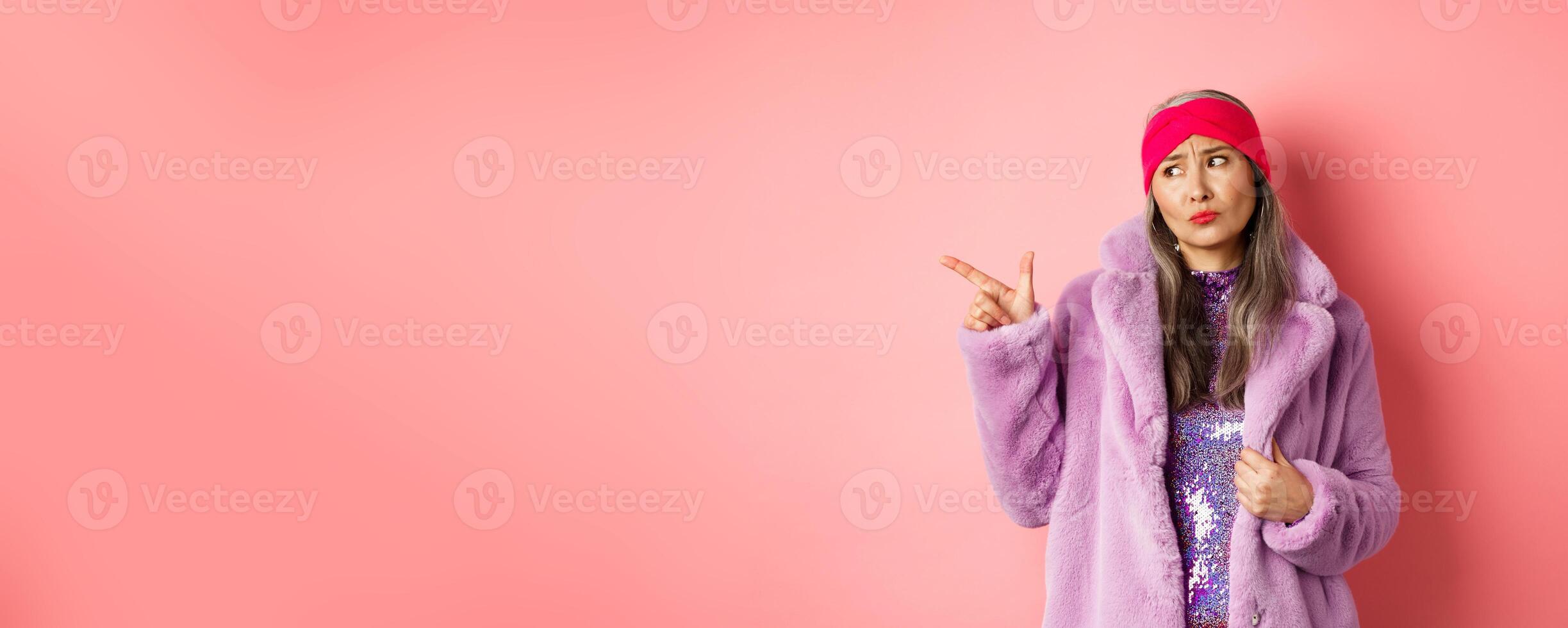 Fashion and shopping concept. Skeptical middle-aged asian woman in purple fur coat looking and pointing finger left with displeased, sad expression, standing over pink background photo