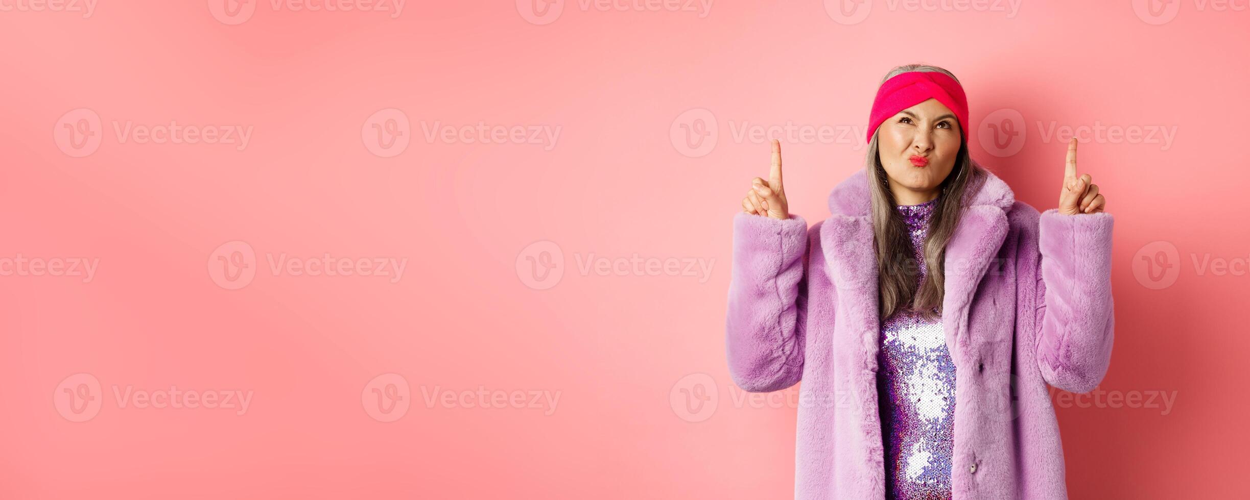 Stylish middle aged asian woman in purple fake fur coat, looking skeptical and grimacing with displeased face, pointing fingers up at something bad and unimpressive, pink background photo