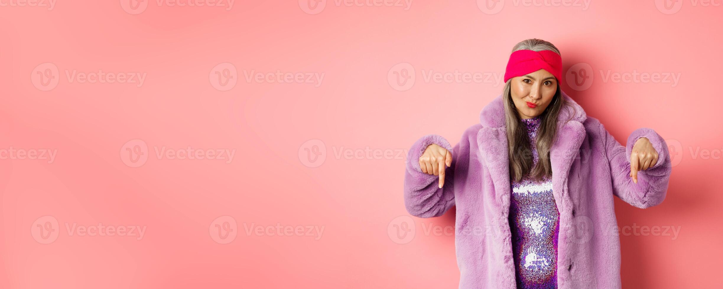 Fashion and shopping concept. Cool asian senior woman pointing fingers down, check out promo offer, looking sassy at camera, pink background photo