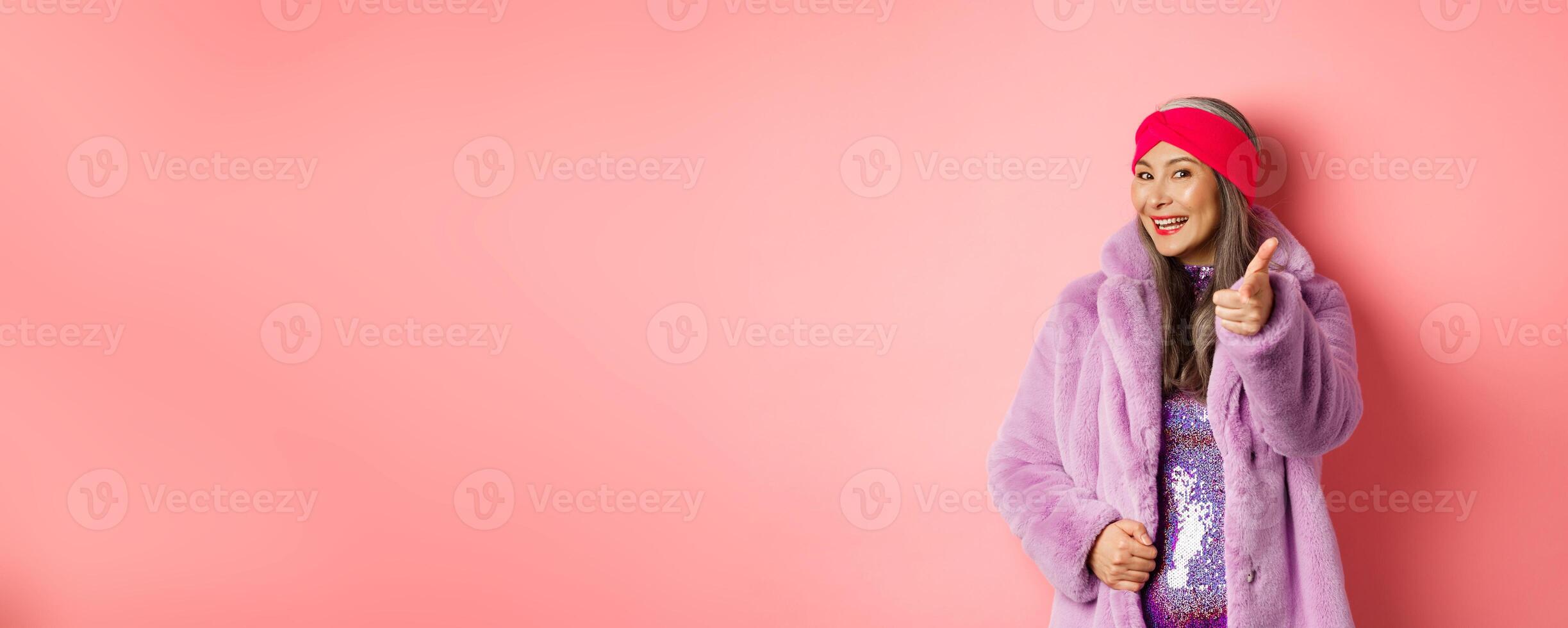 Fashion and shopping concept. Happy asian mature woman in stylish fake-fur coat pointing finger at camera, we need you gesture, standing over pin background photo