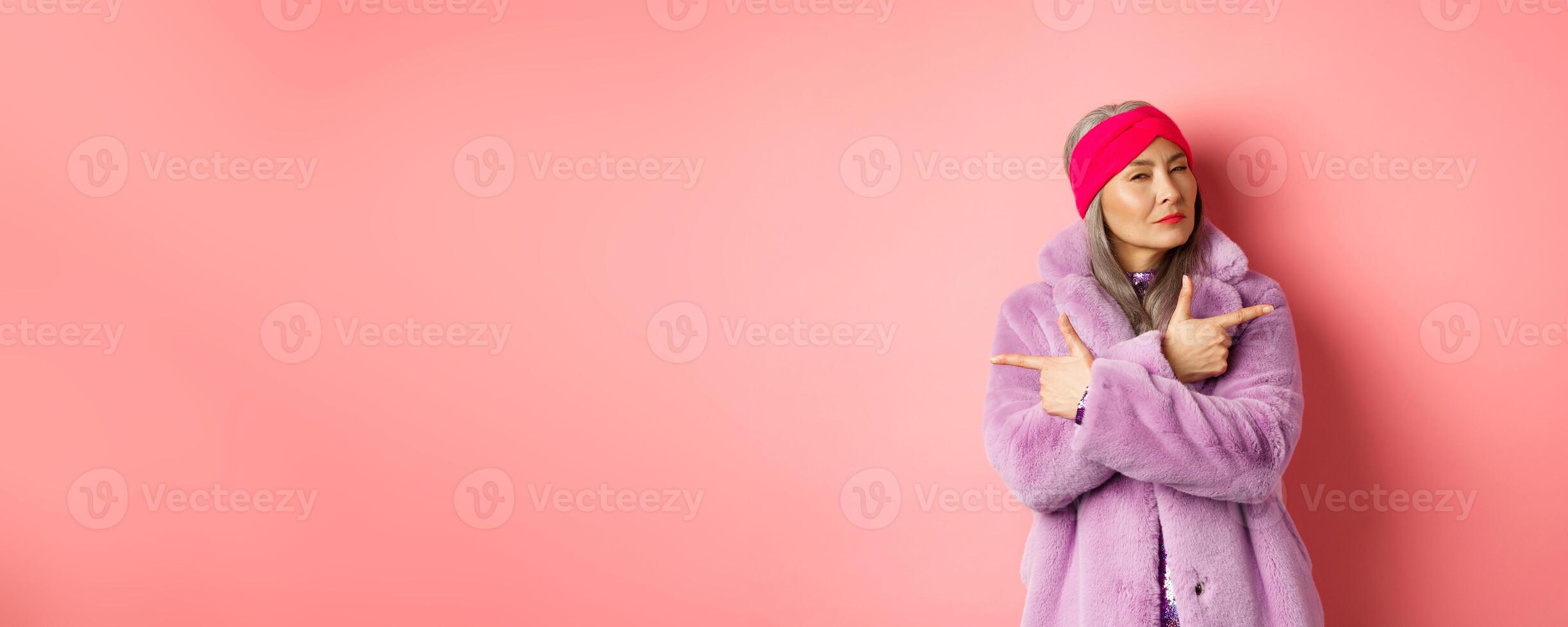 Fashion and shopping concept. Stylish asian mature woman making decision, squinting thoughtful while pointing fingers sideways, showing two variants on pink background photo