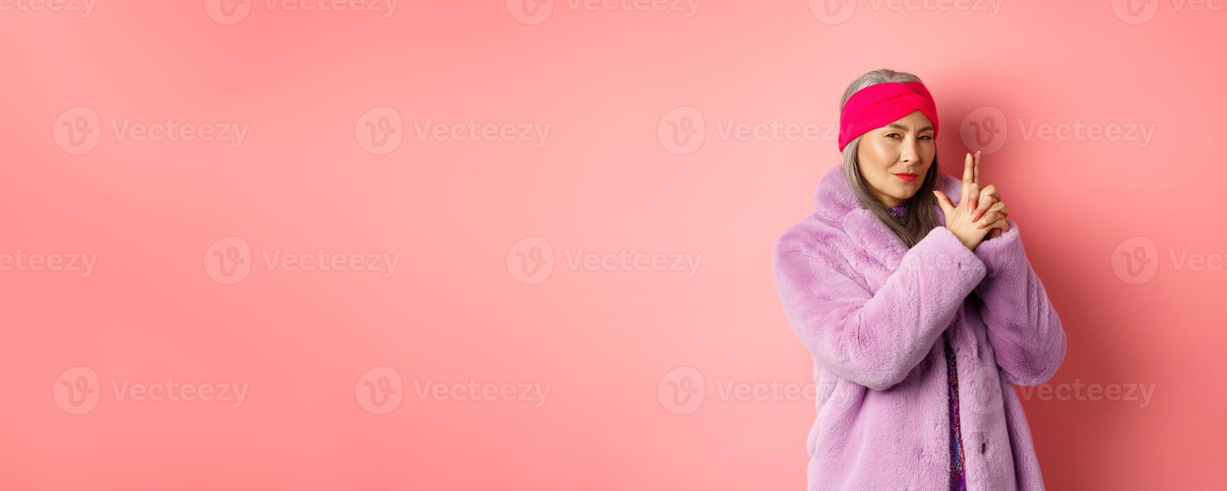Fashion and shopping concept. Cool and stylish asian old woman in winter coat making finger gun gesture and looking sassy at camera, mimicking spy or secret agent with weapon, pink background photo