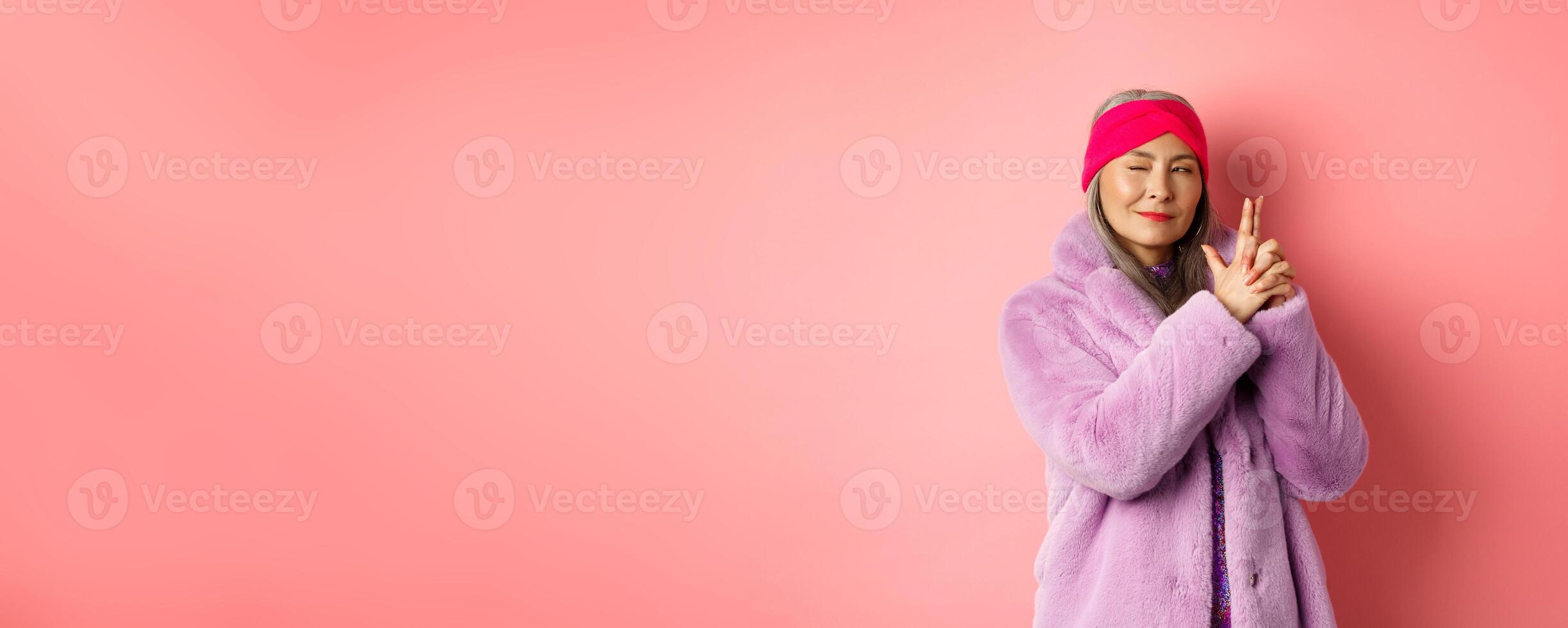 Fashion and shopping concept. Cool and stylish asian senior lady in purple fake fur coat, making finger gun gesture and looking left with sassy smile, acting like secret agent, pink background photo