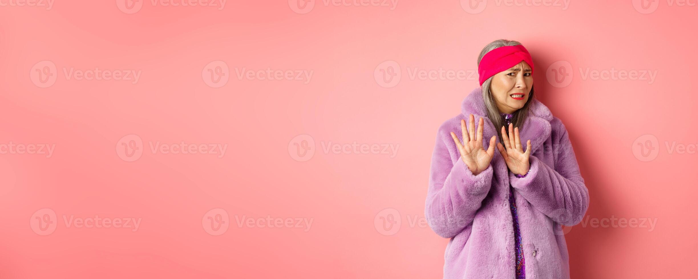 Portrait of stylish asian senior woman in purple winter coat, cringe from something disgusting, frowning and turn away awkward, showing stop gesture, refusing on pink background photo
