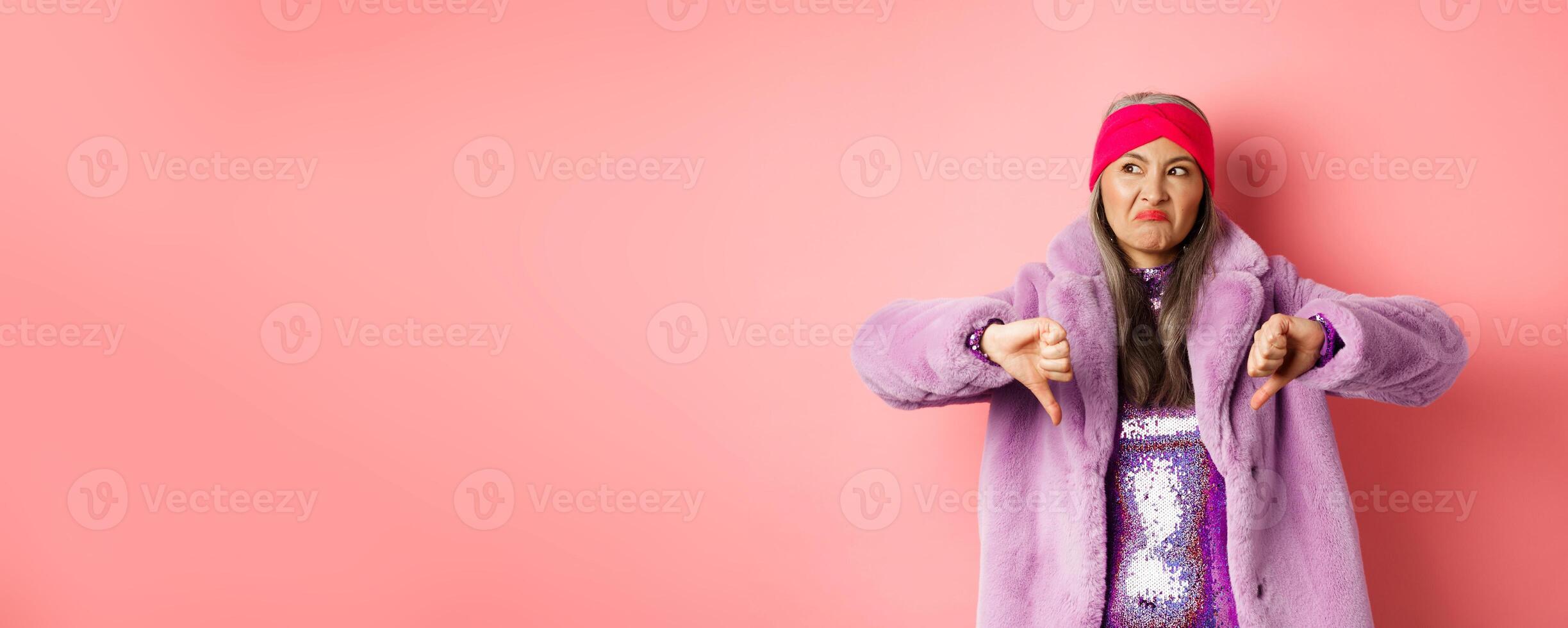 Fashion and shopping concept. Disappointed stylish senior woman showing thumbs-down, looking displeased and judgemental, being dissatisfied, standing over pink background photo
