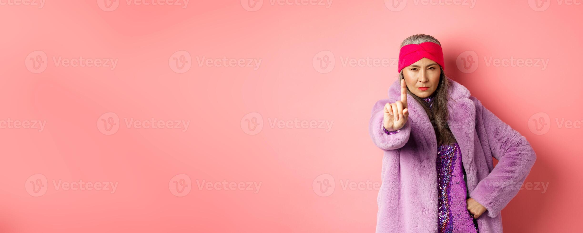 Fashion and shopping concept. Serious asian senior woman showing not so fast gesture, shaking extended finger to stop or warn you, looking determined at camera, wearing stylish purple clothes photo