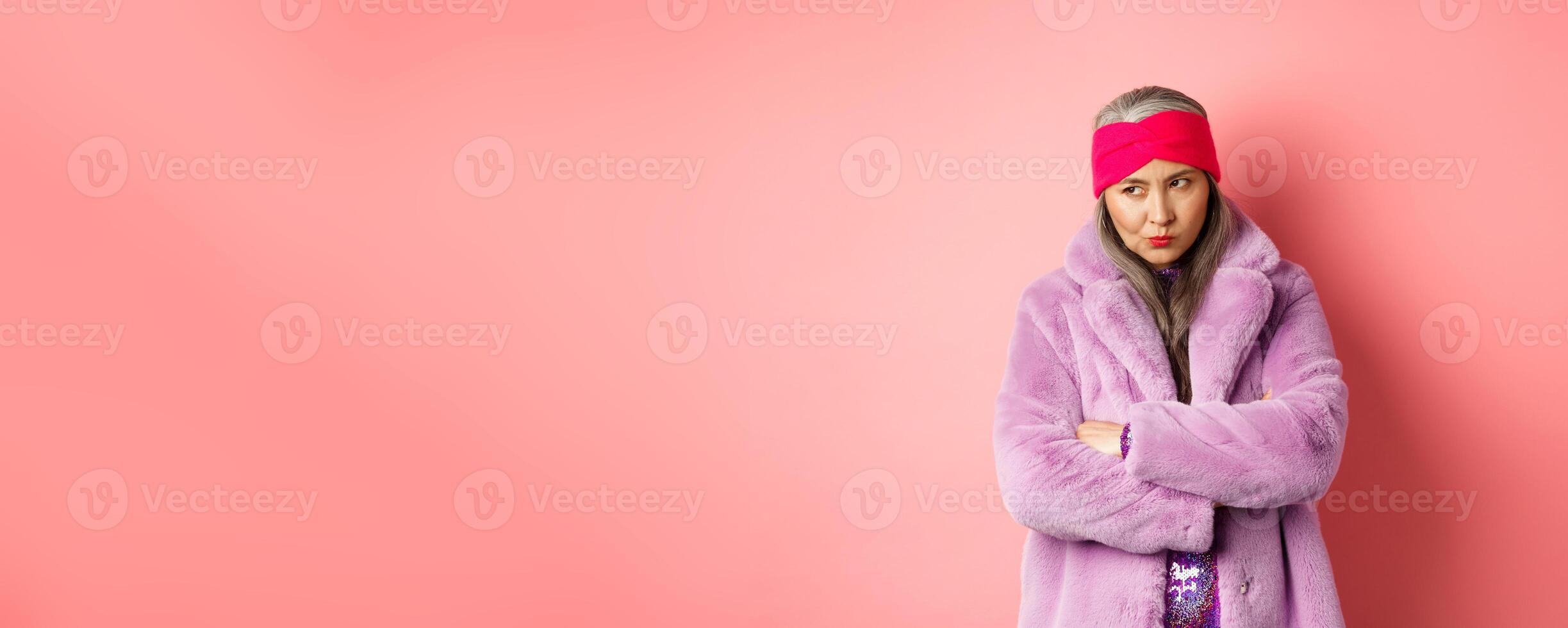 Grumpy funky old asian lady looking from under forehead, pouting and looking left with angry and disappointed face, standing in stylish purple faux fur coat against pink background photo
