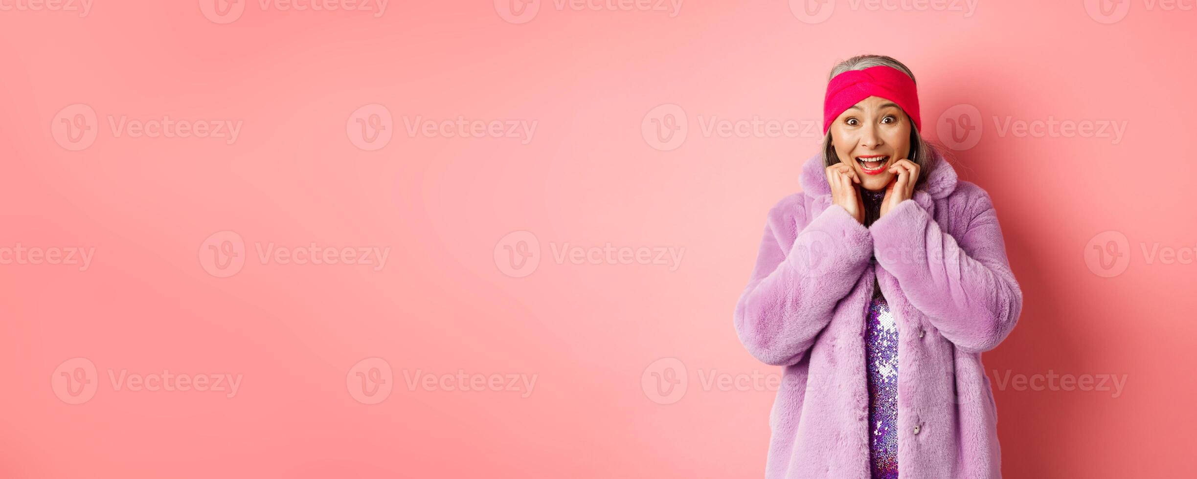 Excited and happy asian female senior model looking at camera with amazement, checking out promo offer, standing in trendy clothes over pink background photo