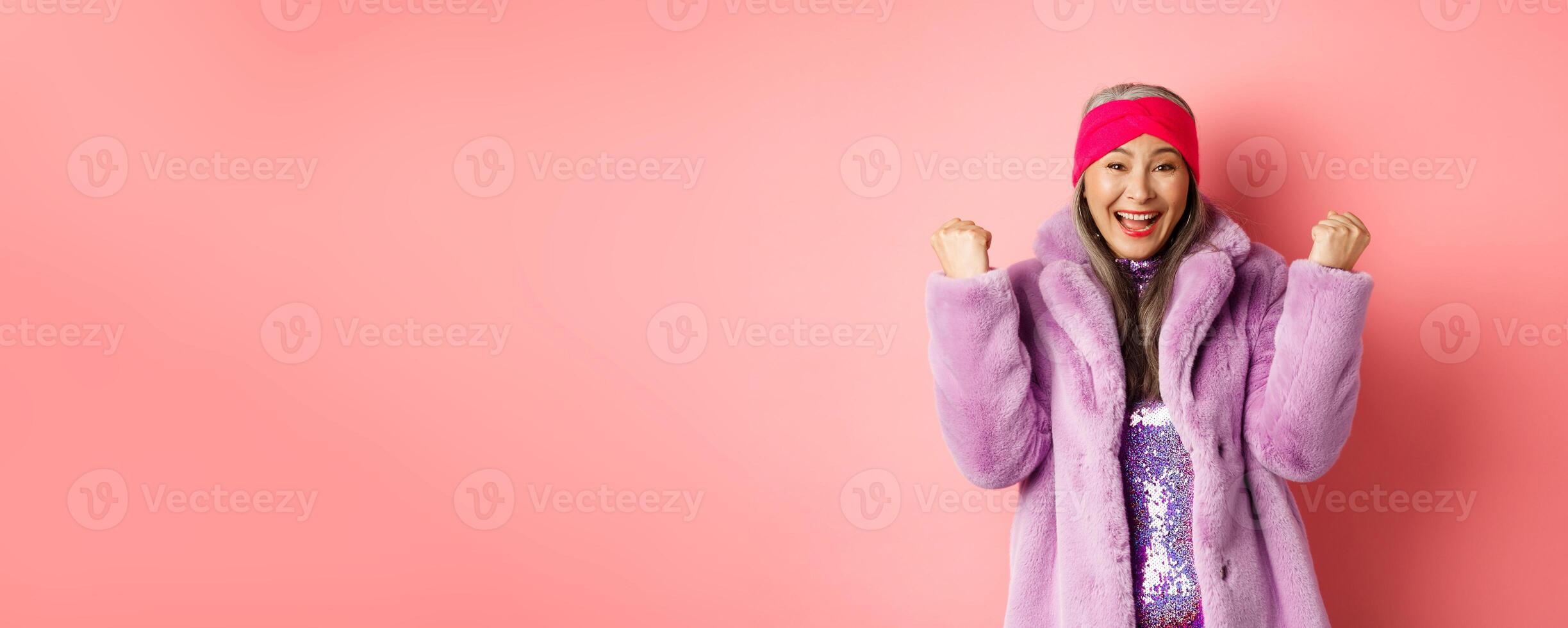 Cheerful asian senior woman winning prize and celebrating. Old funky lady saying yes and clench fists in triumph, standing over pink background photo