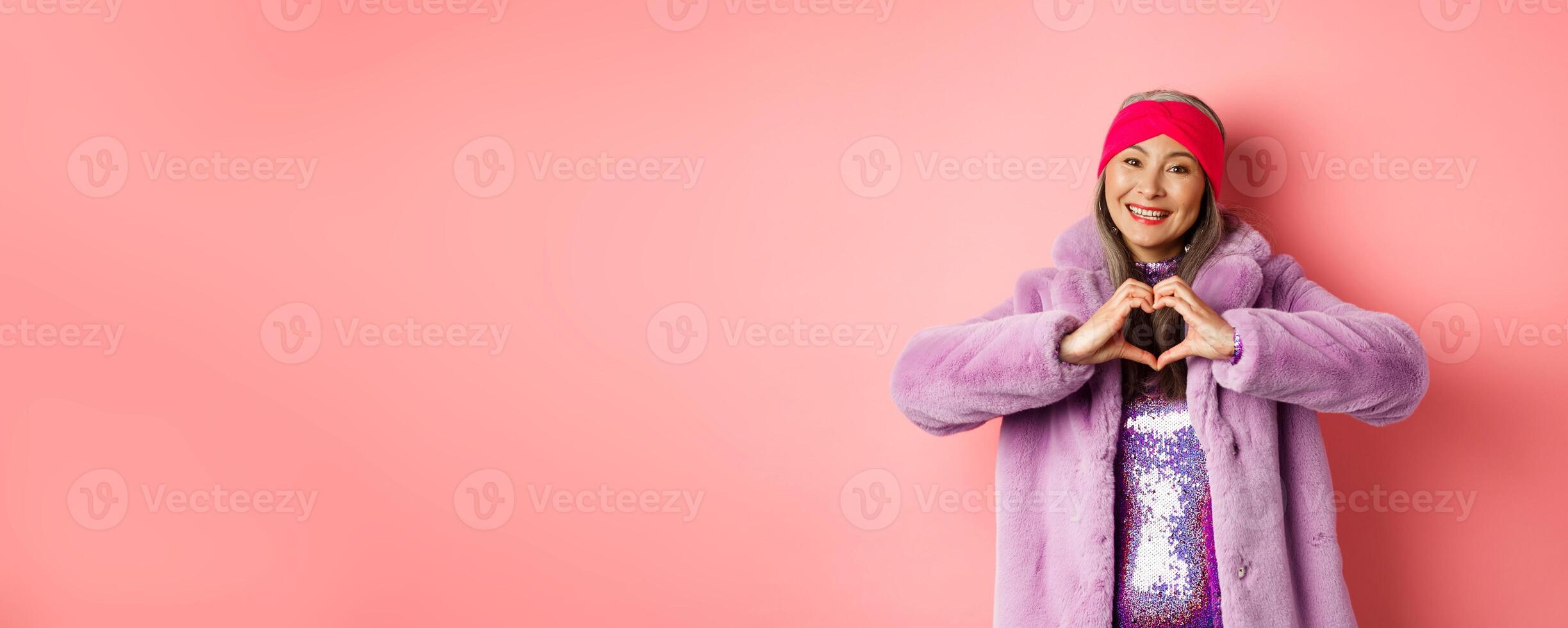 Romance and valentines day. Happy asian senior woman showing heart sign, I love you gesture, smiling and looking caring at camera, standing in faux fur coat, pink background photo