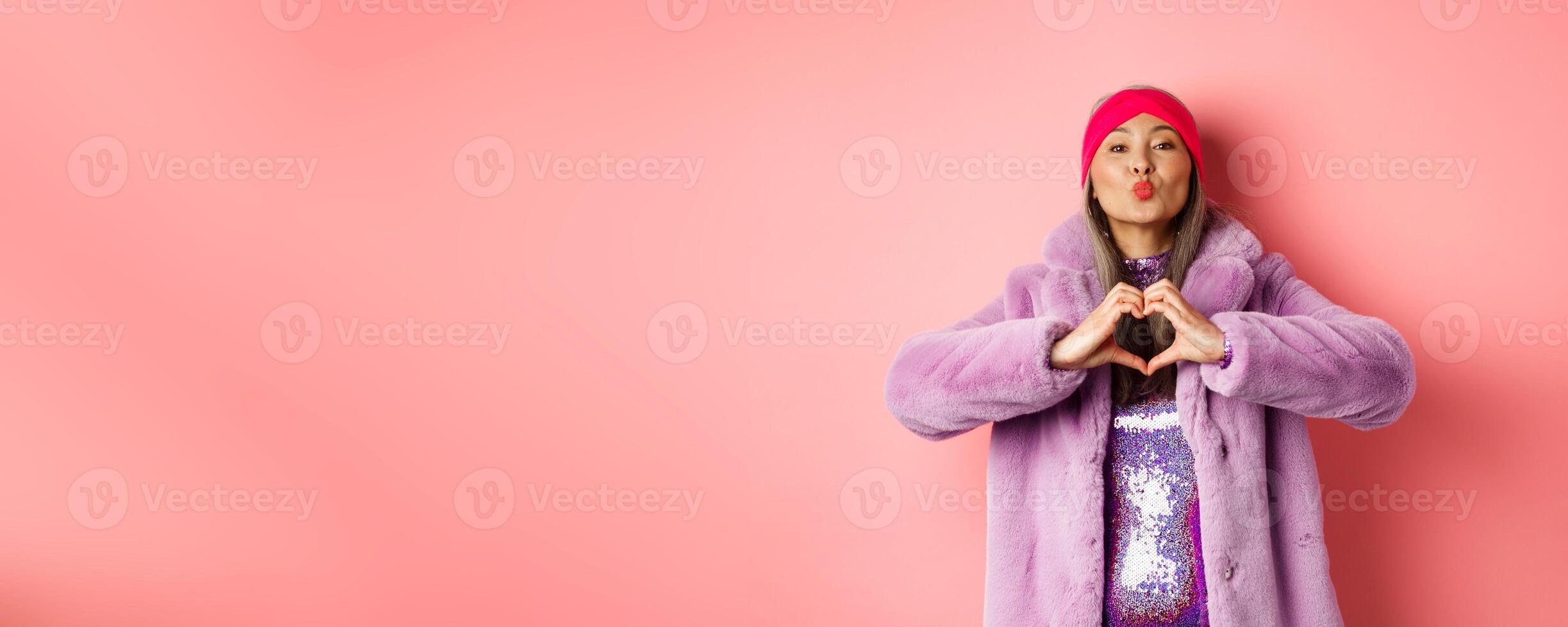 Romance and valentines day. Happy asian senior woman showing heart sign, I love you gesture, pucker lips for kiss, standing over pink background photo