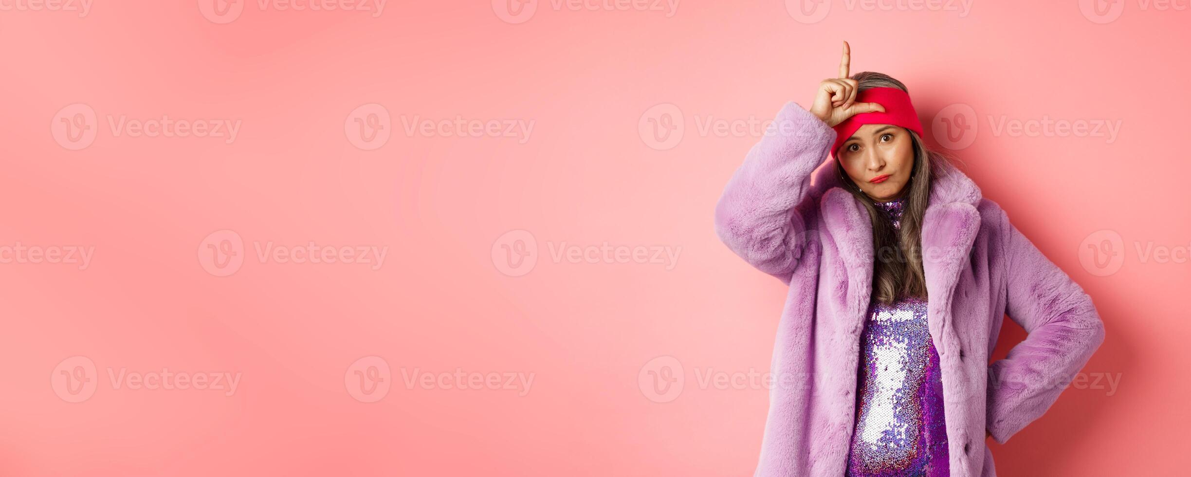 Stylish and cool asian mature woman showing loser sign on forehead, mocking you, standing in trendy faux fur coat and glittering dress, pink background photo