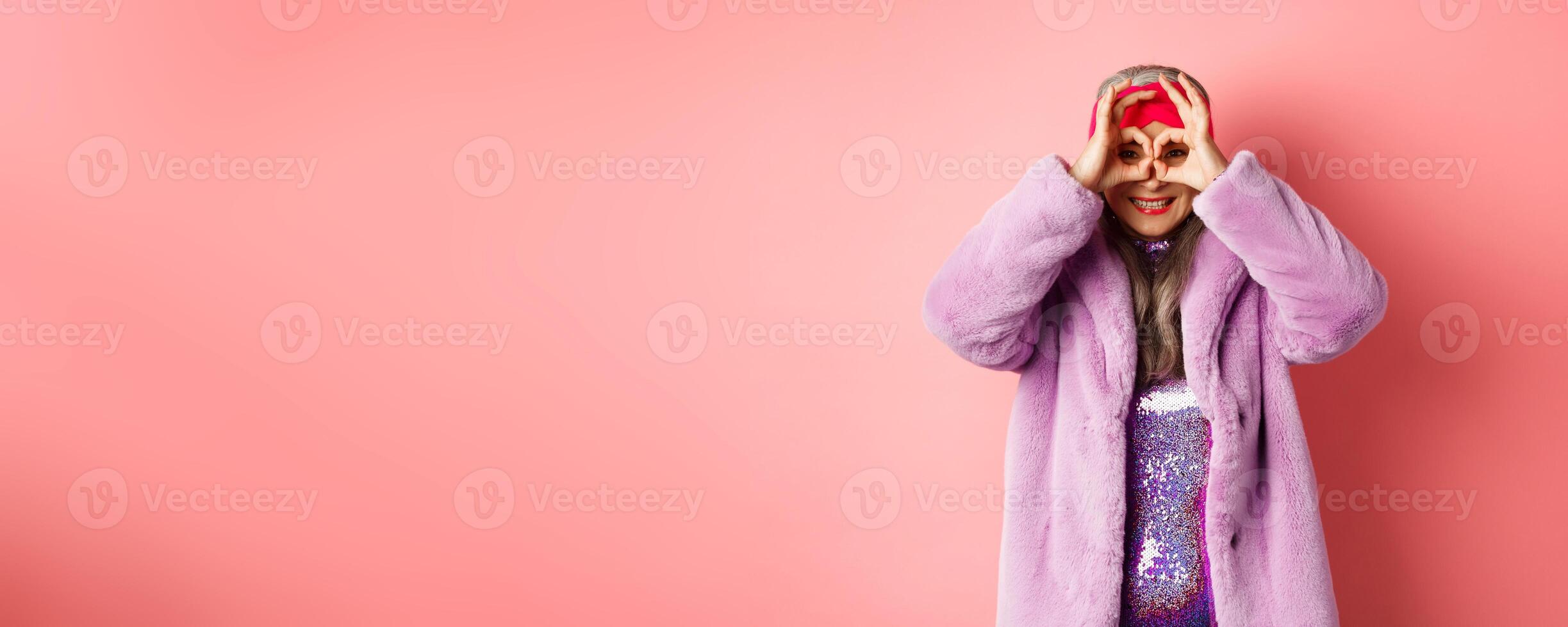 Fashion and shopping. Happy asian grandmother in stylish faur fur coat, looking through hand glasses, making binoculars and smilinga t camera, standing over pink background photo
