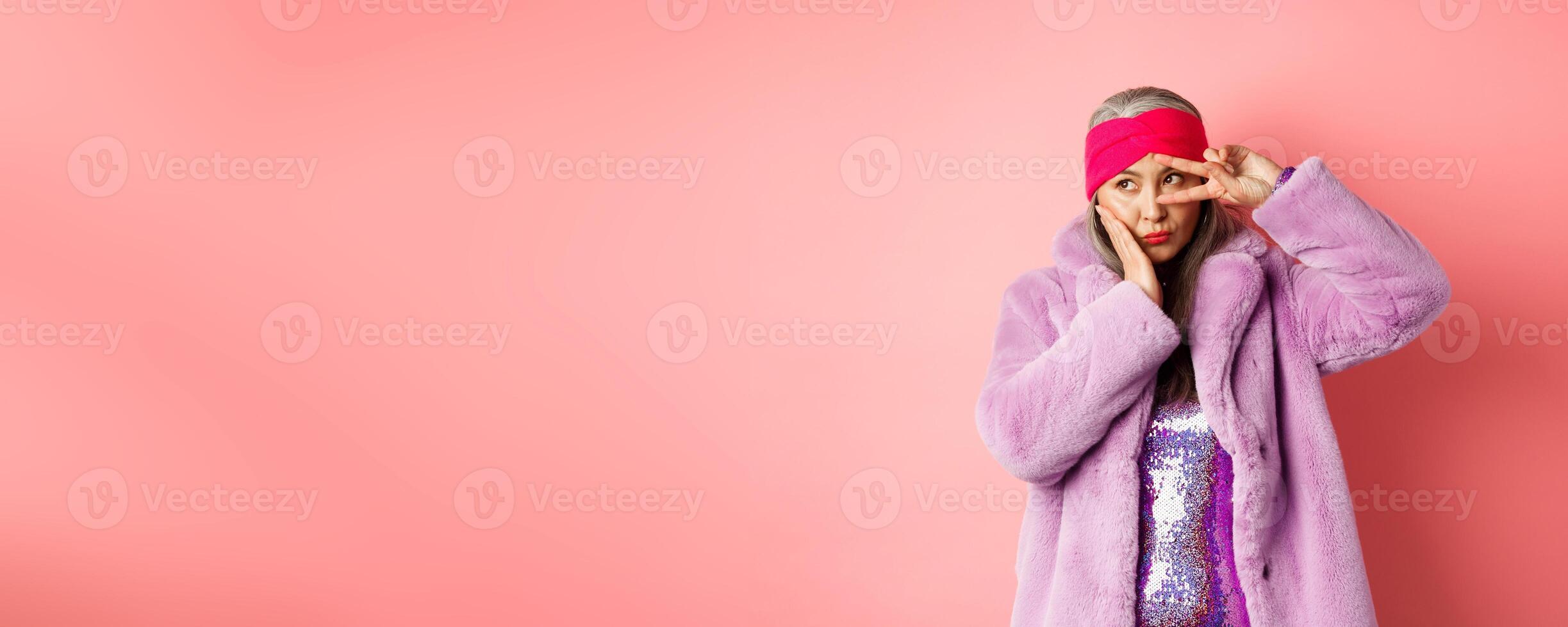 Fashion and shopping. Stylish and funky asian female model, showing peace signs and looking cute and silly, posing in purple faux fur coat over pink background photo