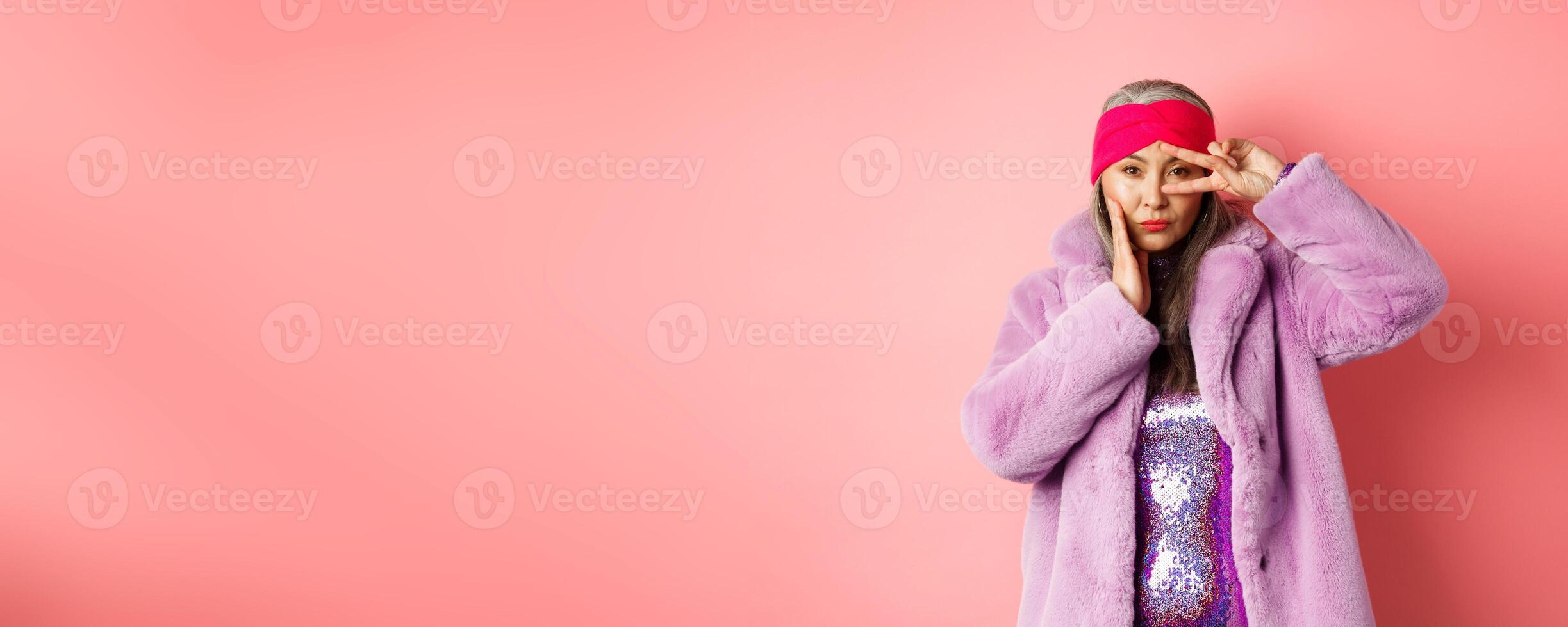 Fashion and shopping. Beautiful asian senior woman in stylish faux fur coat and headband, making peace sign on face and looking sassy and confident at camera, pink background photo