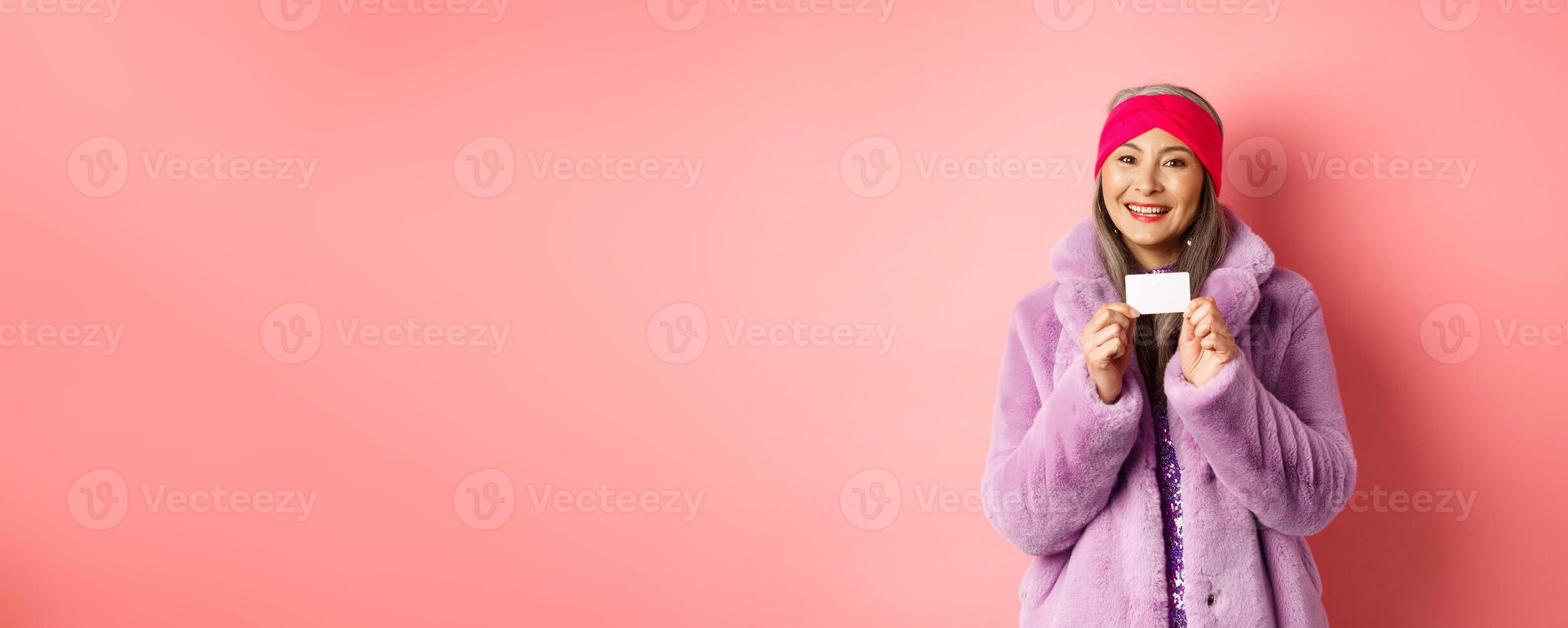 Shopping and fashion concept. Beautiful asian middle-aged woman showing plastic credit card and smiling happy in stylish clothes, pink background photo