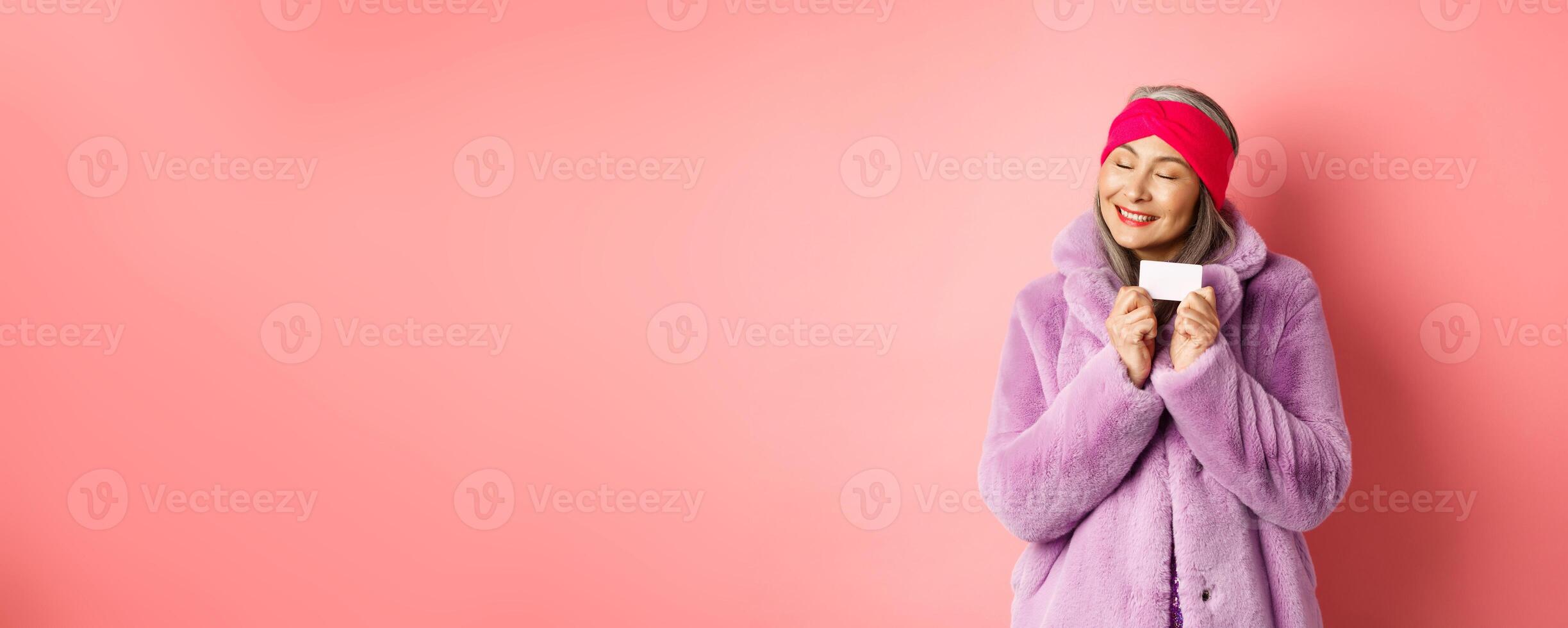 Shopping and fashion concept. Fashionable asian woman in purple faux fur coat, looking happy and showing plastic credit card, smiling cheerful, standing over pink background photo