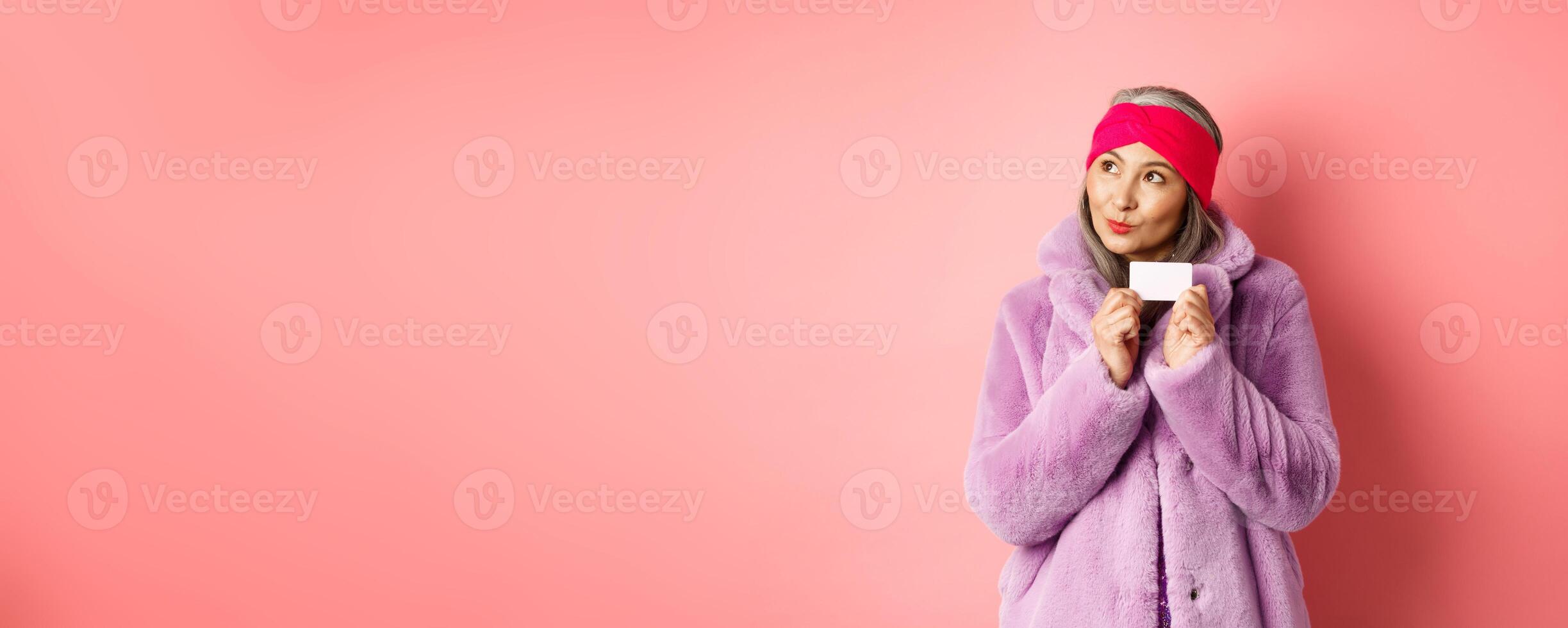Shopping and fashion concept. Fashionable asian senior woman thinking about buying gifts, holding credit card and looking thoughtful at upper left corner, pink background photo