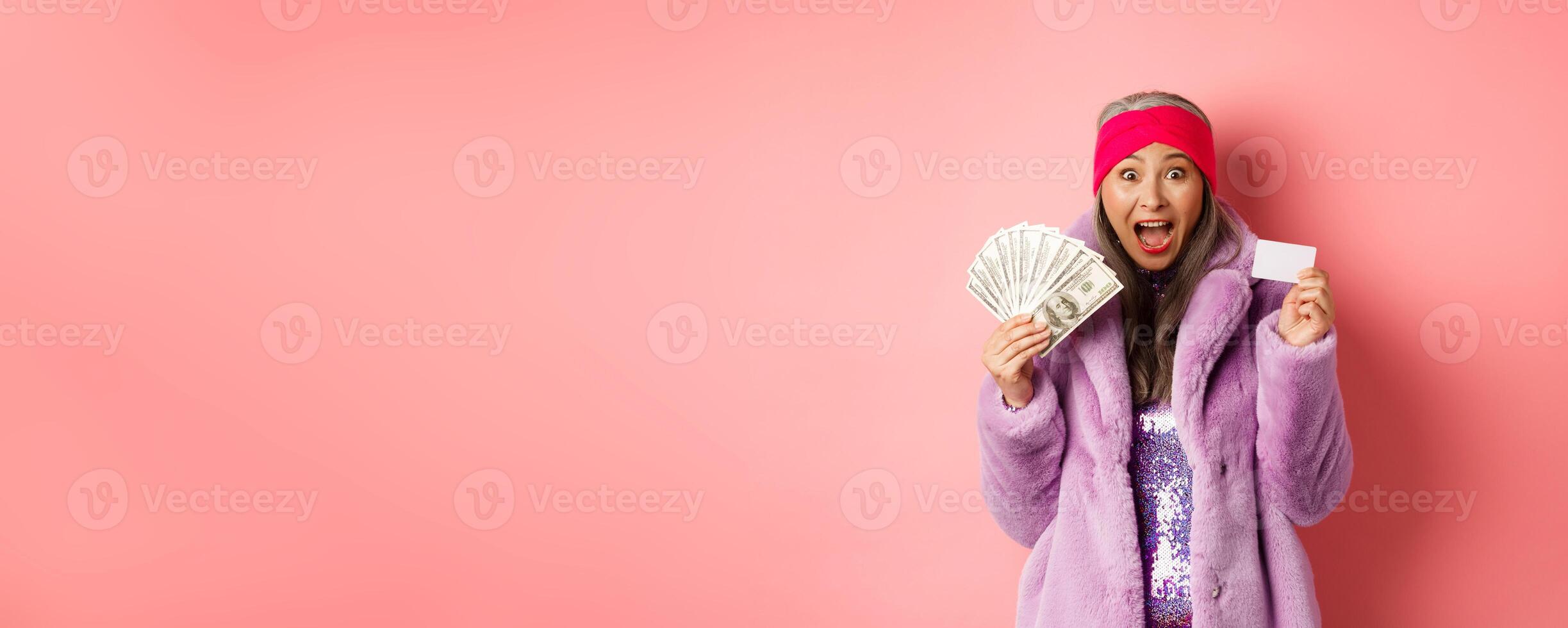 Shopping and fashion concept. Excited asian senior woman going buy something with money and plastic credit card, scream of joy and happiness, pink background photo