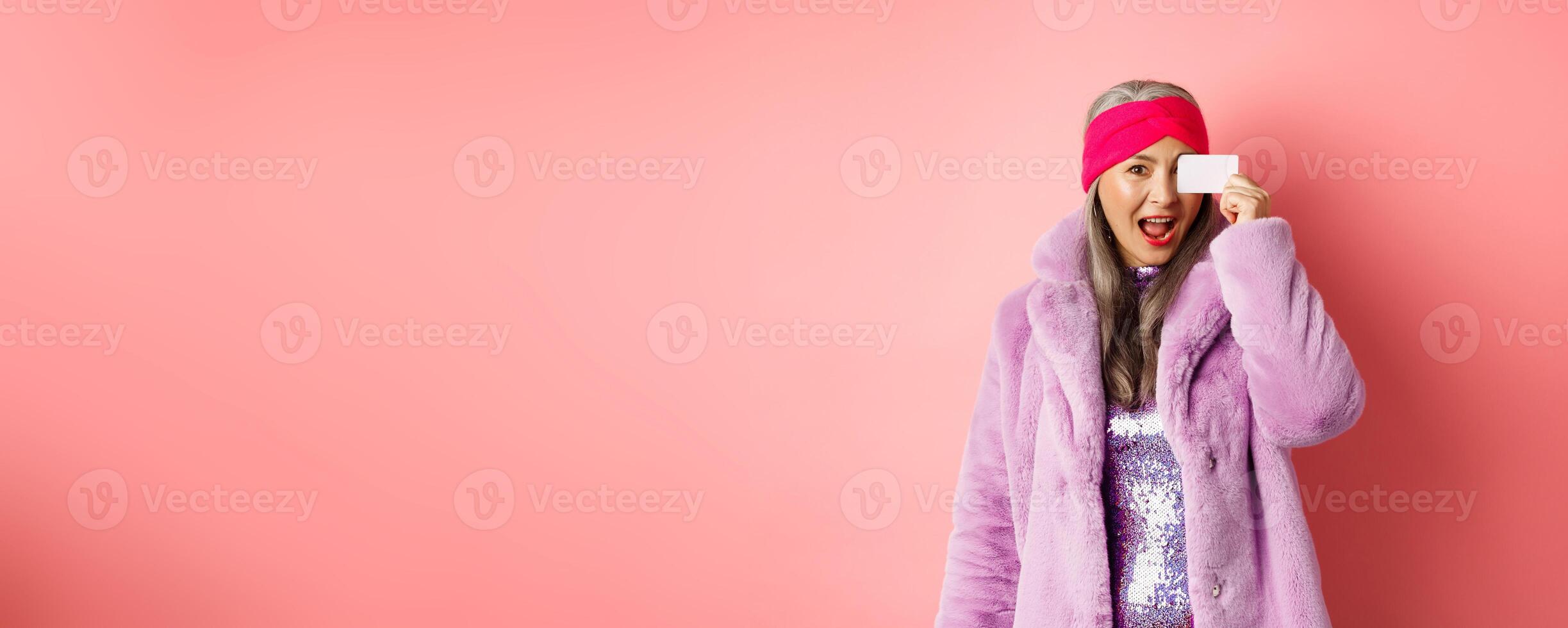 Shopping and fashion concept. Beautiful asian middle-aged woman showing plastic credit card on face and looking excited at camera, standing over pink background photo