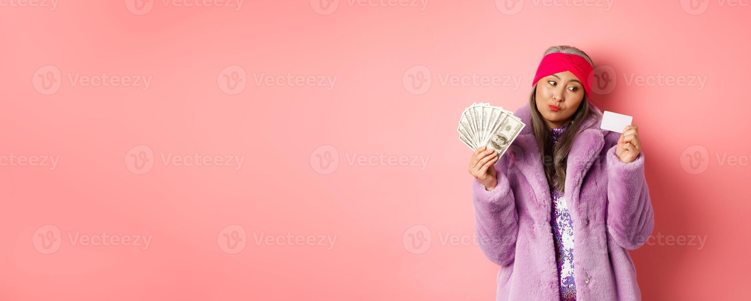 Shopping and fashion concept. Thoughtful asian female holding dollars but thinking about paying contactless with plastic credit card photo