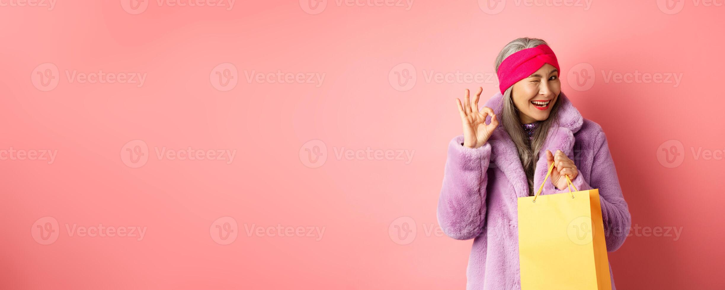 Stylish senior asian woman recommending store, holding shopping bag and winking with OK sign, smiling satisfied, standing over pink background photo