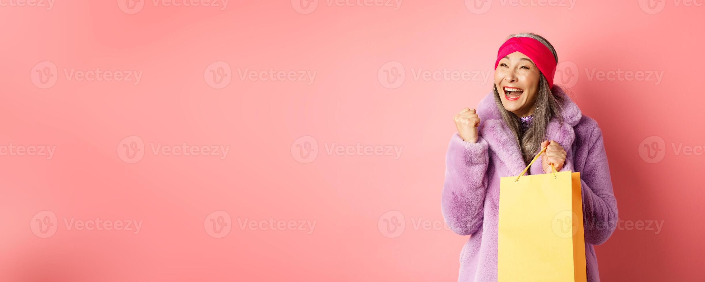 Shopping and fashion concept. Happy asian senior woman winning, holding paper bag and making fist pump gesture, saying yes with cheerful face, standing over pink background photo