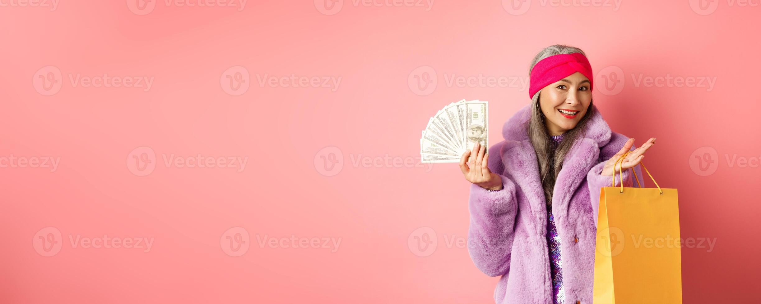 Rich and stylish grandmother going on shopping with dollars, holding paper bag and smiling carefree, standing over pink backgrund photo