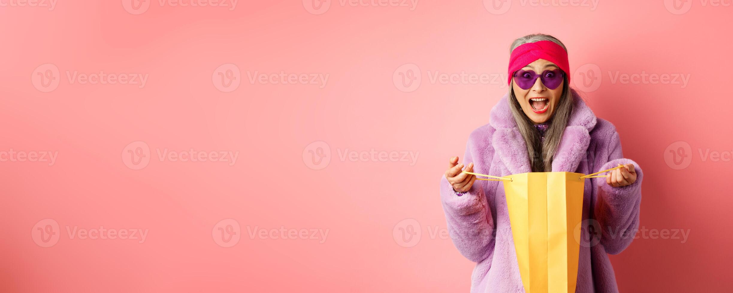 Shopping and fashion concept. Stylish asian elderly woman in sunglasses and faux fur coat open paper bag with gifts, looking amazed at camera, pink background photo