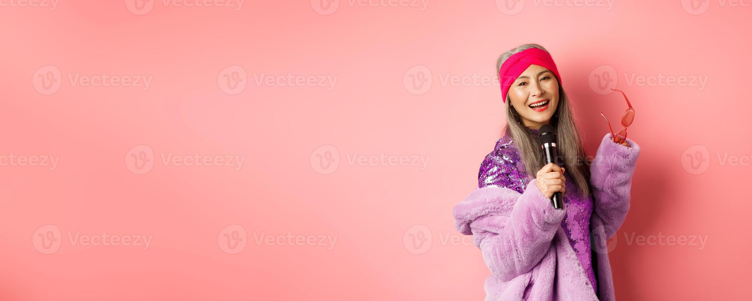 Gorgeous asian senior woman singing karaoke in mic, perform song and looking happy, standing over pink background photo