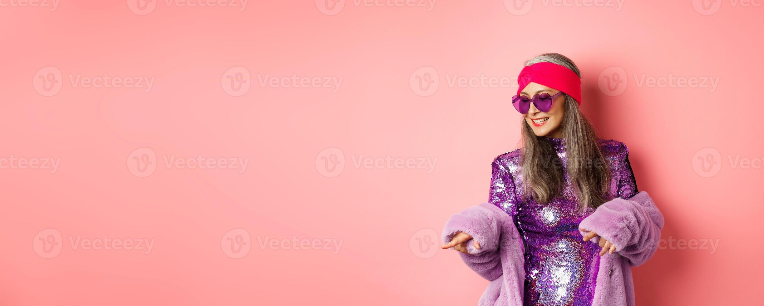 Fashionable asian senior woman dancing disco, wearing glittering shiny dress and heart-shaped sunglasses, having fun, standing over pink background photo